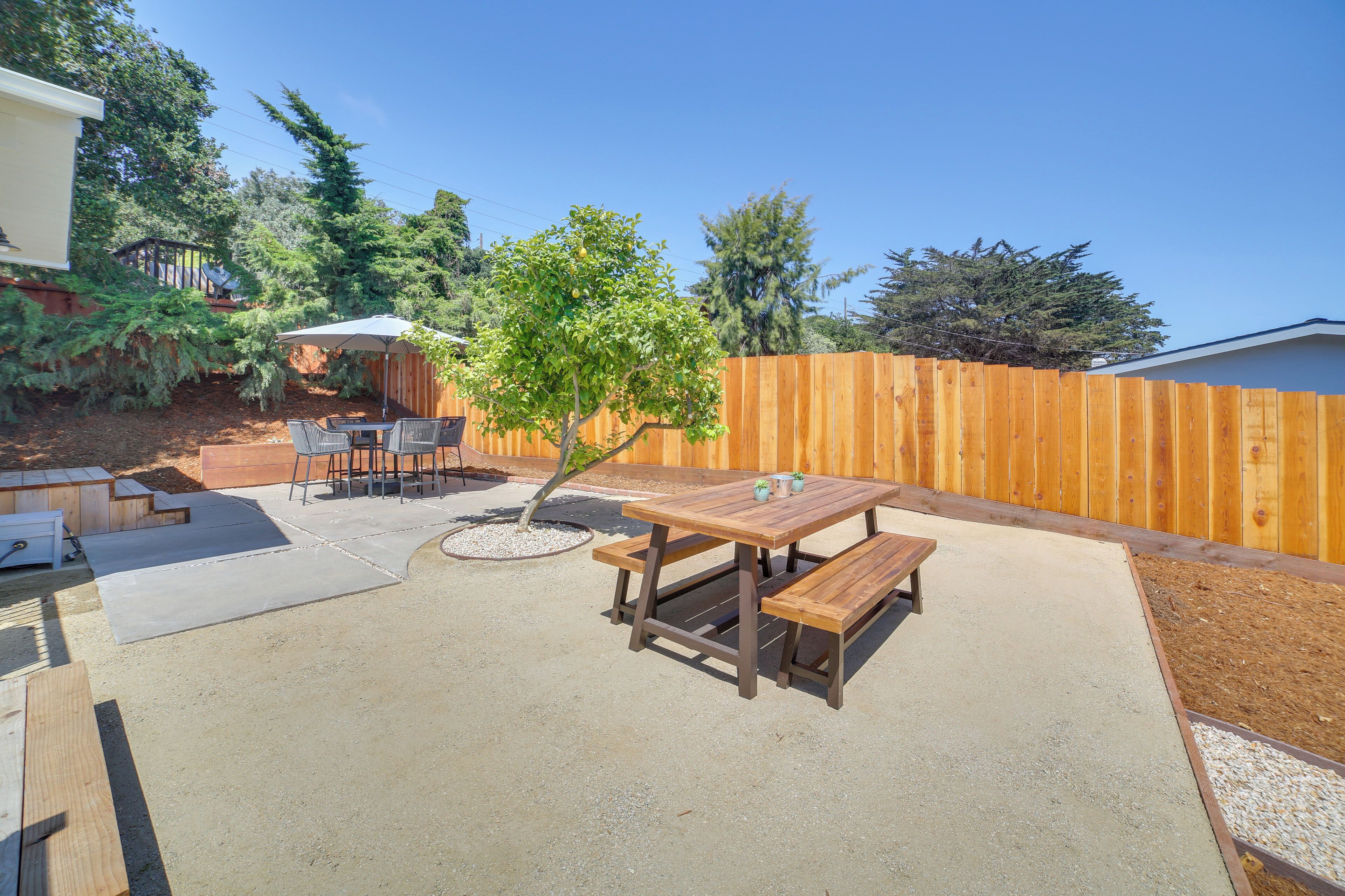 Une terrasse à l’arrière de la maison, avec une table et des bancs en bois, entourée d’une clôture en bois et de différentes plantes.