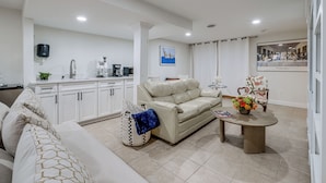 A modern living room with a beige sofa, a coffee table, and a dining table. There is a kitchen area with white cabinets and a black coffee machine.