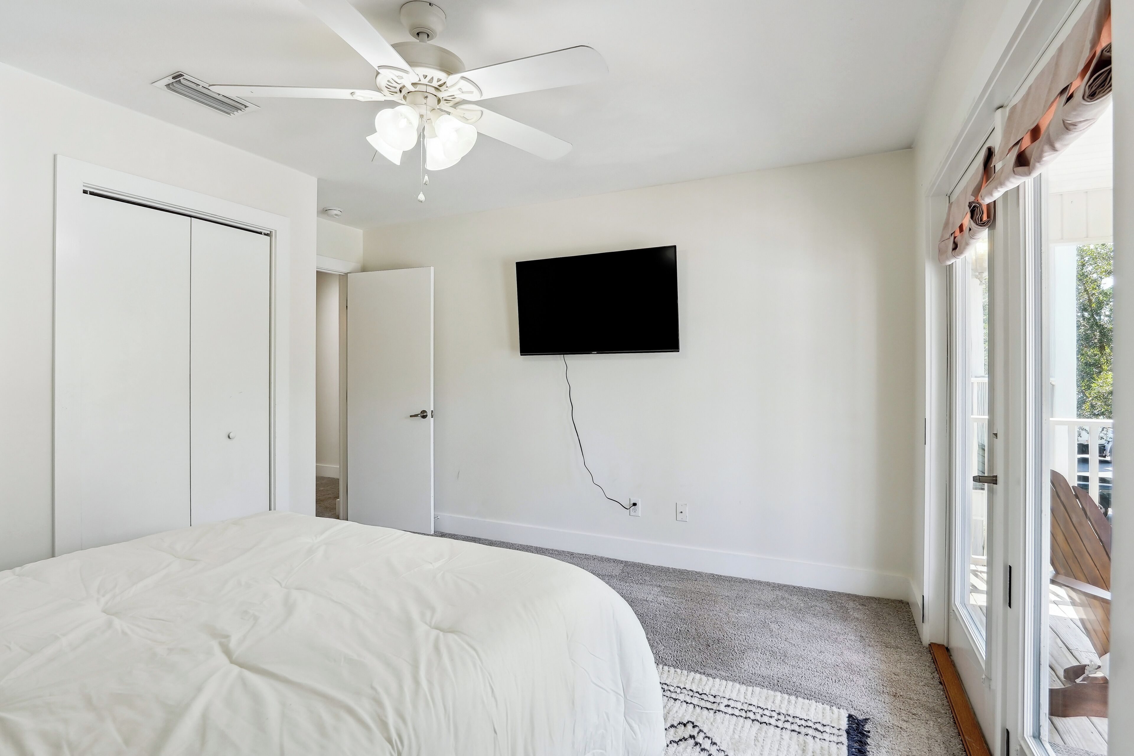 Un dormitorio con cama, ventilador de techo, televisor montado y puerta corrediza de vidrio al exterior.