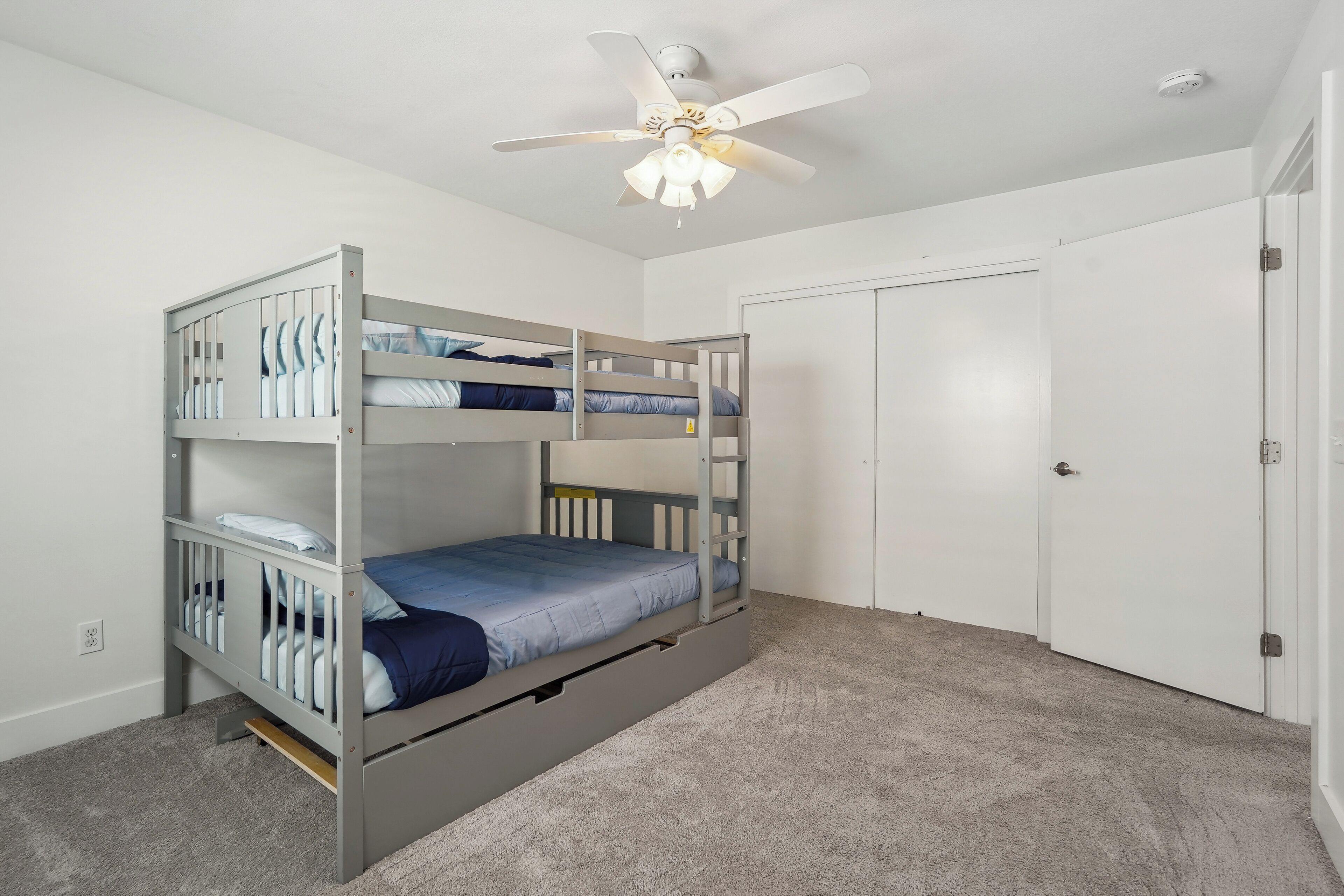Camera con letto a castello, ventilatore a soffitto, pareti bianche e pavimento rivestito a moquette.