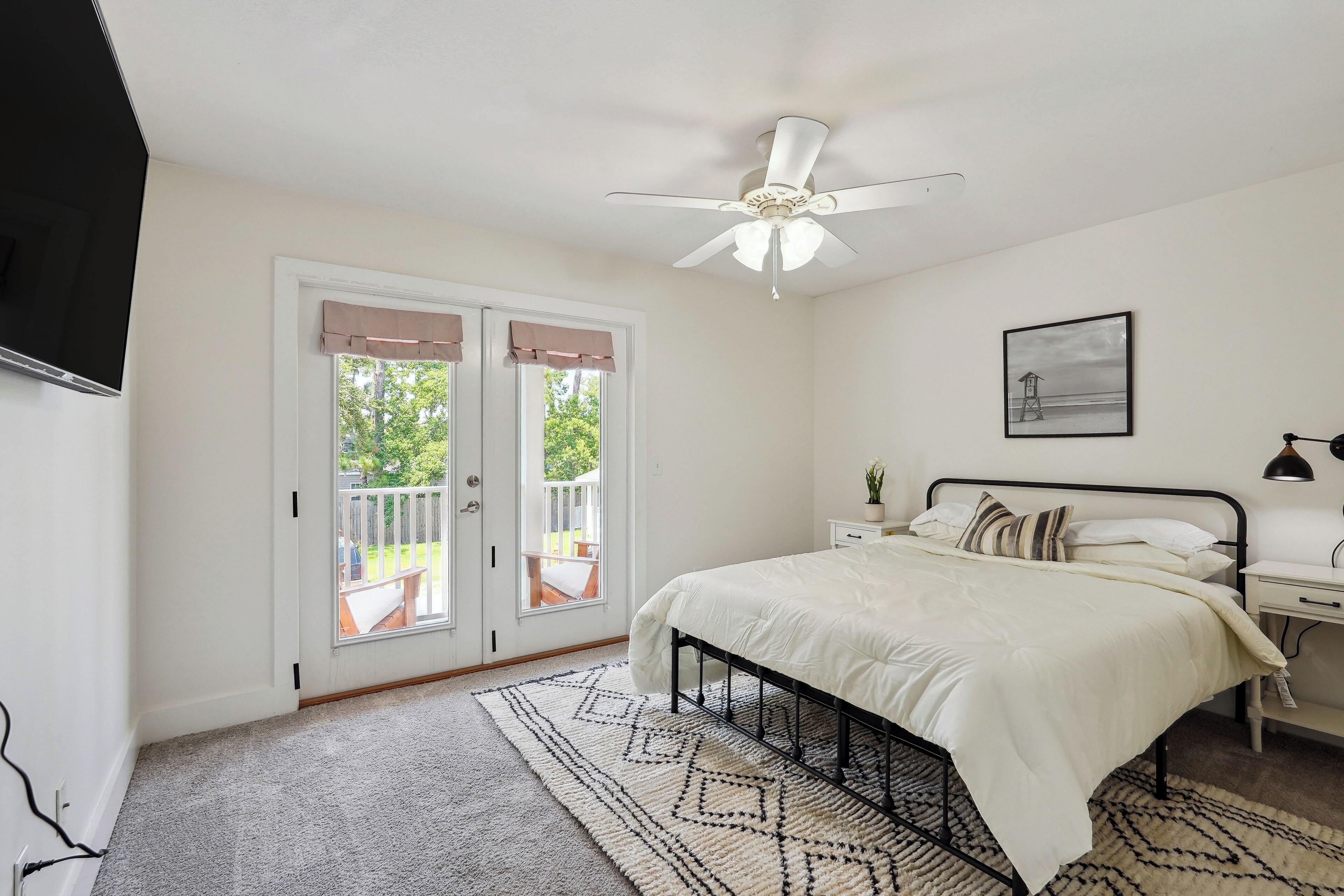 A bedroom with a bed, bedside table, nightstand, ceiling fan, and a view through a sliding door to an outdoor balcony.