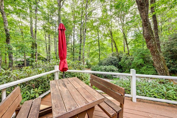 Une terrasse en bois avec une table et des bancs, entourée d’une végétation luxuriante et d’arbres.