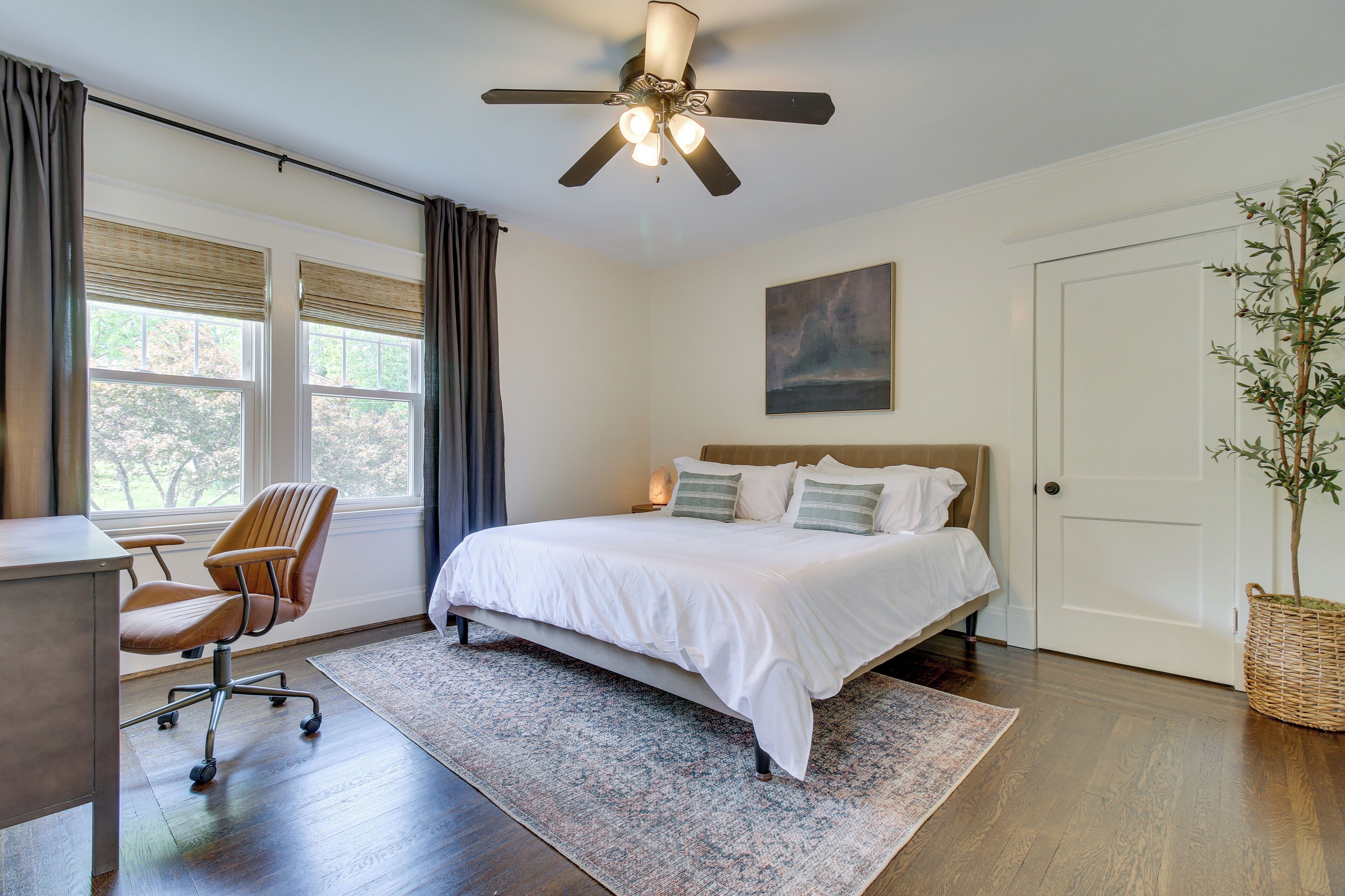 A bedroom with a bed, a desk, a chair, a ceiling fan, a window with curtains, and a door.