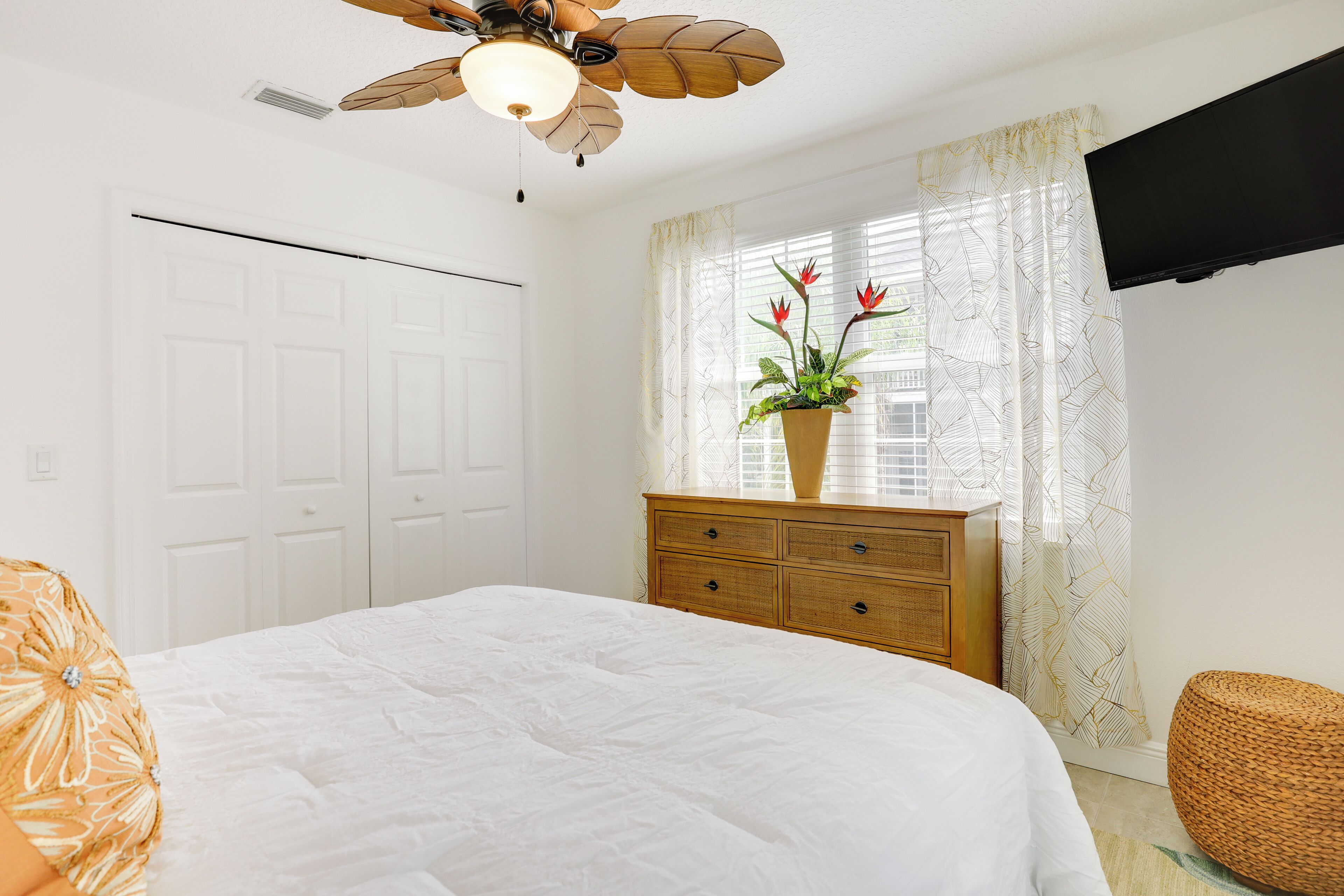 A bedroom with a bed, a wooden dresser, a ceiling fan, a window with curtains, and a television on the wall.