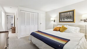 A hotel room with a bed, white walls, a framed picture, and a lamp.