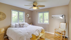 A bedroom with a large bed, two windows, a ceiling fan, a bedside table with a lamp, and a rocking chair.
