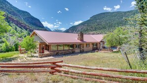 A lodge with a stone facade, large windows, and a red roof, surrounded by mountains and a wooden fence. - Beautiful Ouray Home w/ Patio - 3 Mi to Downtown! (Ouray)