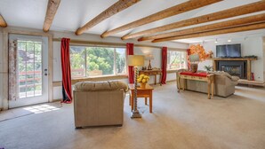 A spacious living room with a sofa, a table with a lamp, and a television. There are large windows with curtains, a fireplace, and exposed wooden beams on the ceiling. - Beautiful Ouray Home w/ Patio - 3 Mi to Downtown! (Ouray)