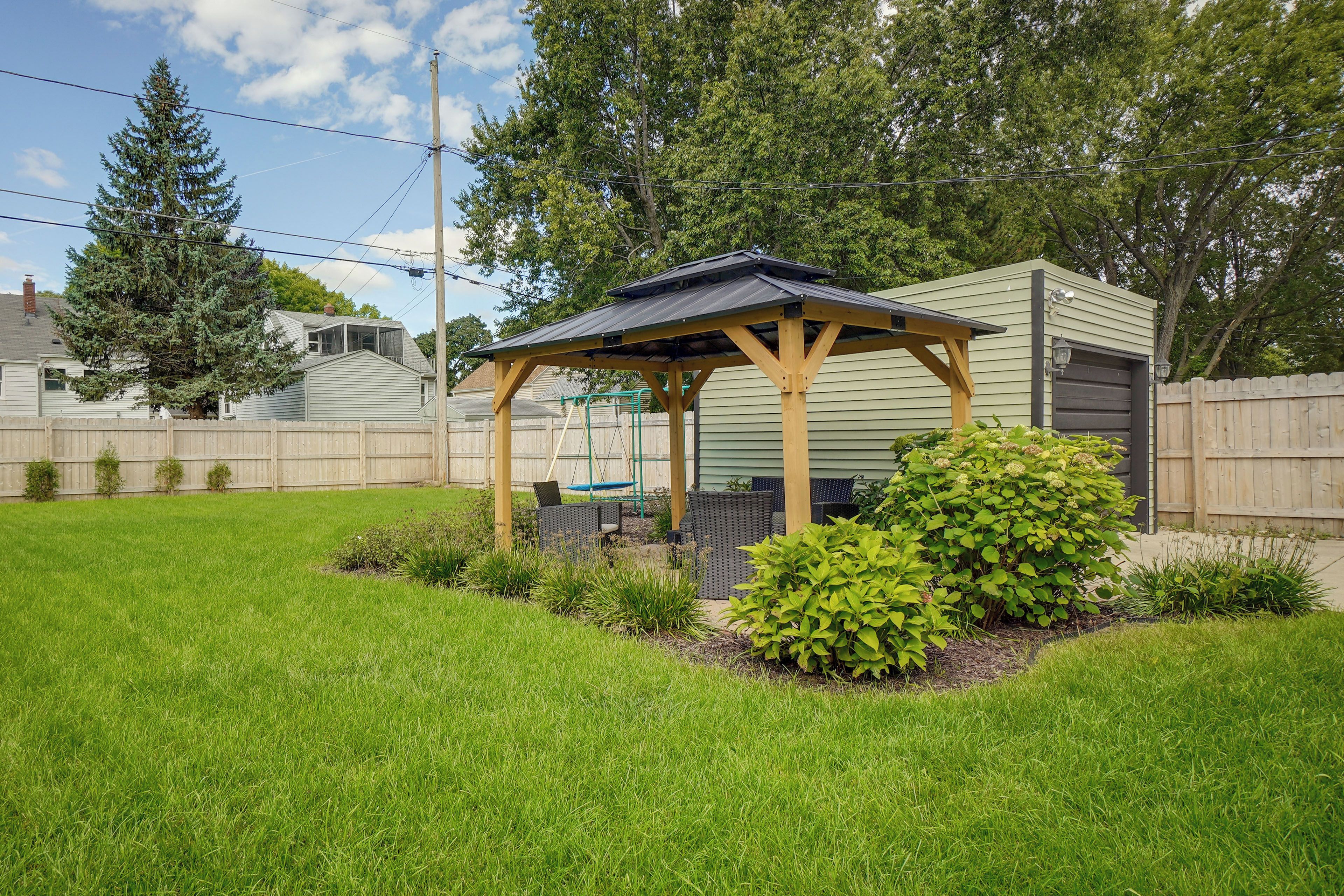 A covered outdoor seating area with a pergola structure, surrounded by a well-maintained lawn and various shrubs.