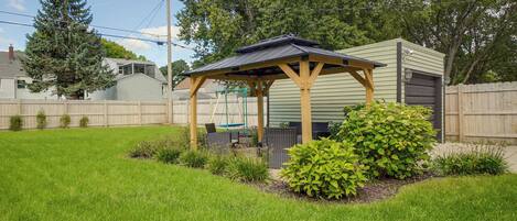 A covered outdoor seating area with a pergola structure, surrounded by a well-maintained lawn and various shrubs.