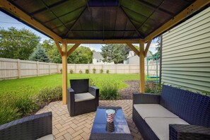 A covered patio area with wicker furniture and a table, surrounded by a wooden fence and a grassy yard. - Milwaukee Home w/ Serene Patio & Backyard Garden! (Milwaukee)