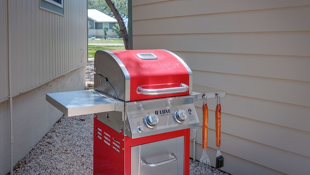 Un barbecue à gaz O’Luxe rouge et argenté, doté de deux boutons de contrôle et d’une étagère latérale, est placé devant une maison dont le revêtement extérieur est de couleur beige.