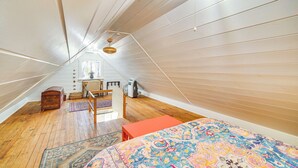 Habitación con techo inclinado, piso de madera, cama con colcha colorida, un pequeño cofre de madera y ventana con cortinas.