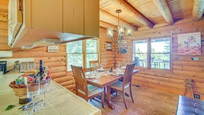 A wooden cabin with a dining area featuring a table set for four, wooden chairs, and a chandelier. - Crestline Area Vacation Rental Cabin w/ Decks! (Cedarpines Park)