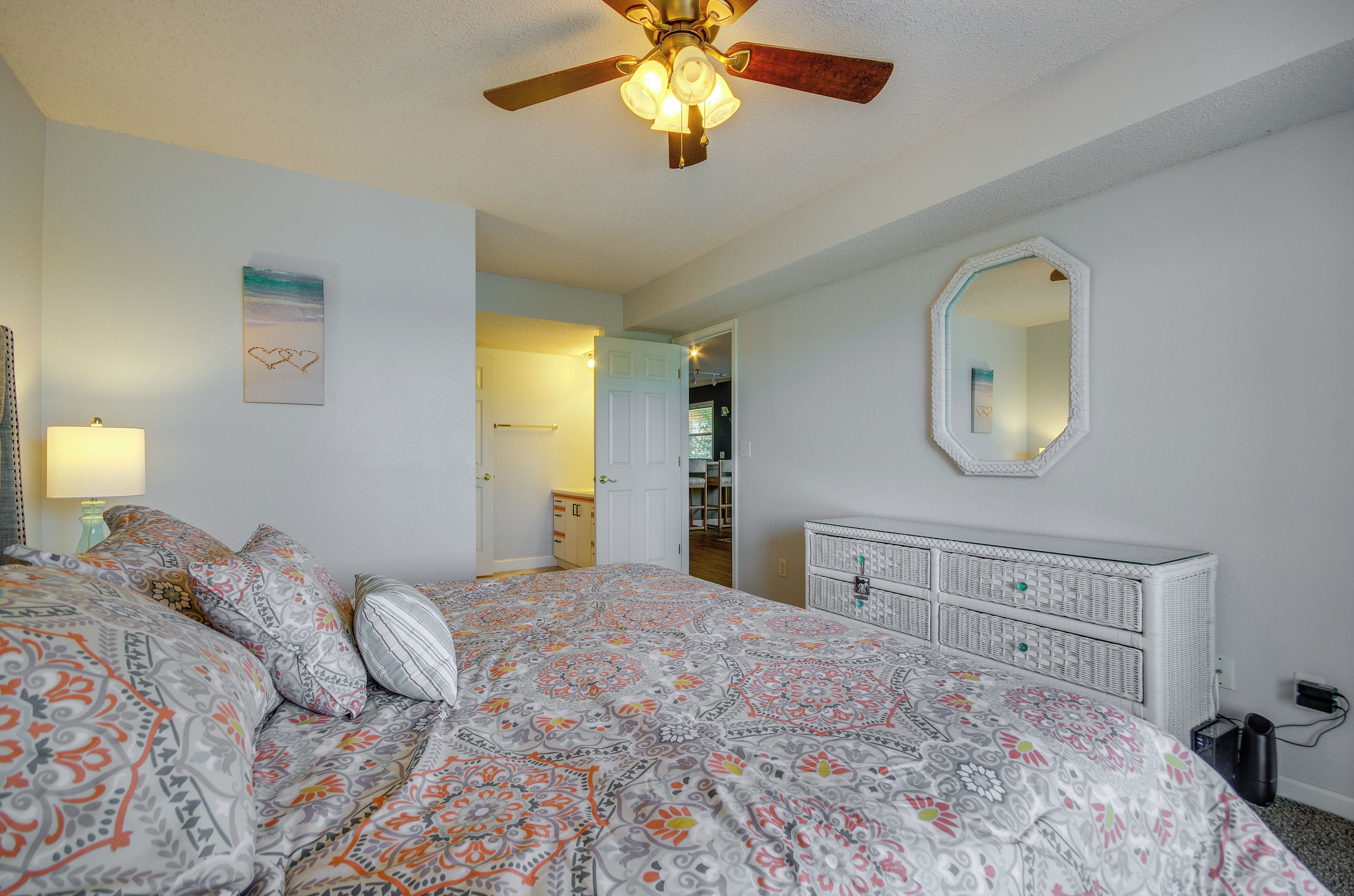 Un dormitorio con cama, ventilador de techo, cómoda, espejo y puerta que da a otra habitación.