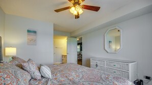 Une chambre à coucher comprenant un lit, un ventilateur de plafond, une commode, un miroir et une porte donnant sur une autre pièce.