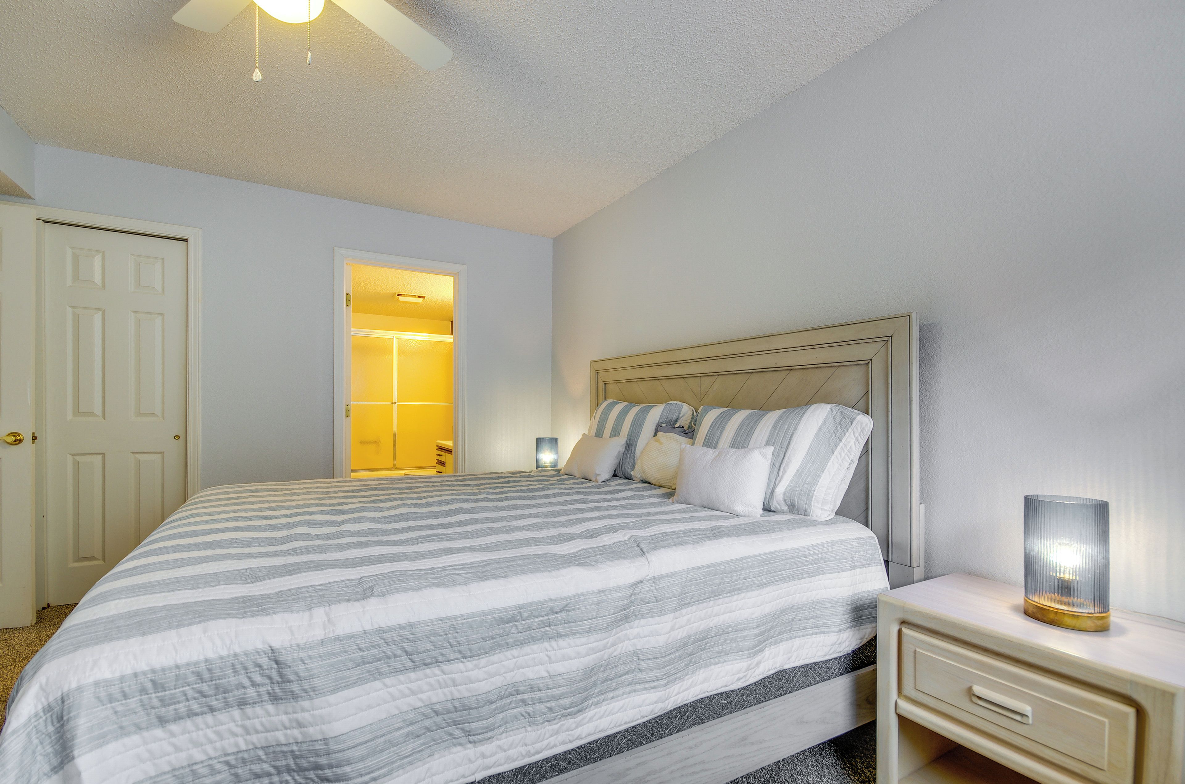 A neatly made bed with striped bedding, a bedside table with a lamp, and a door leading to a bathroom.