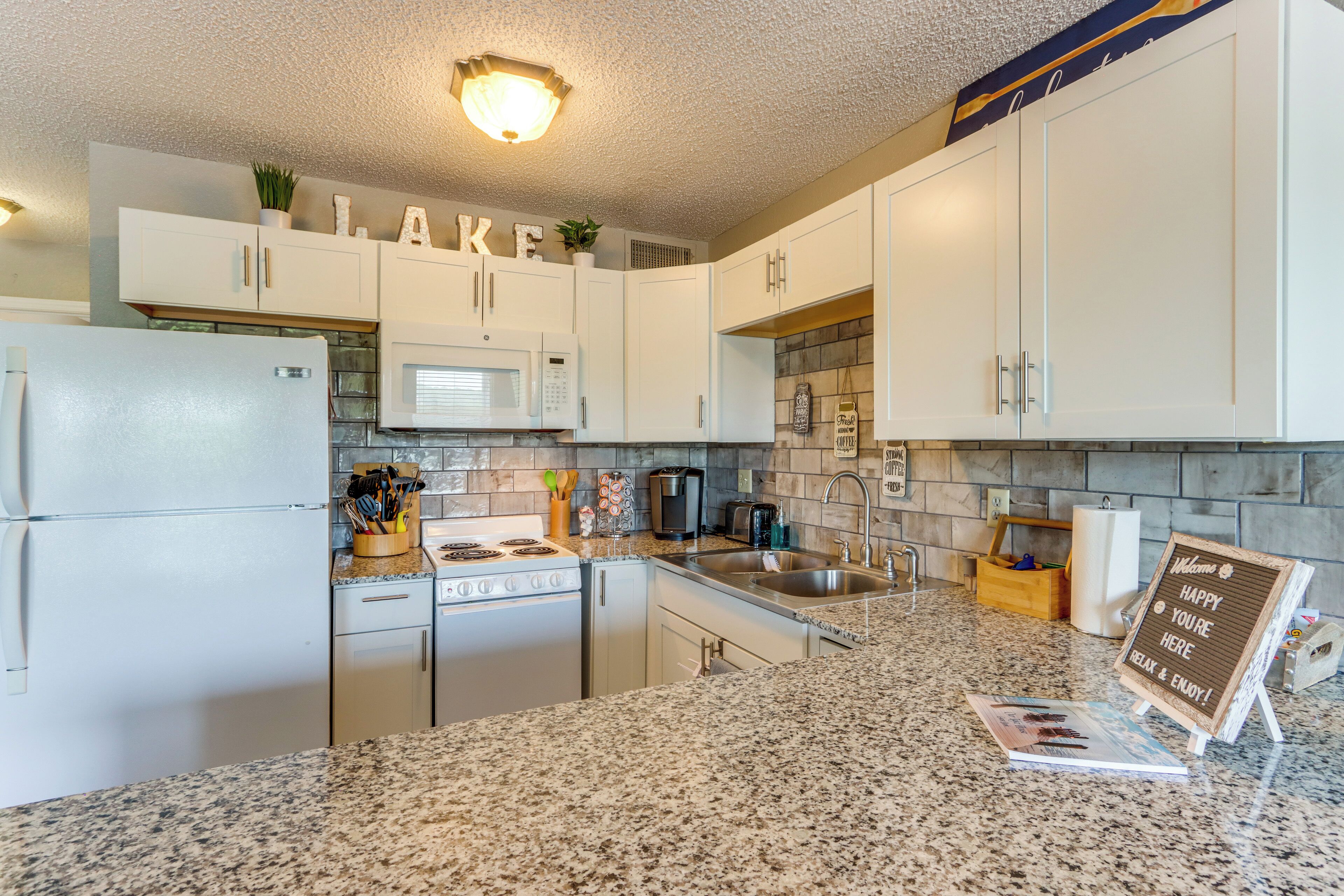 A kitchen with white cabinets, granite countertops, a refrigerator, microwave, stove, sink, and a welcome sign.