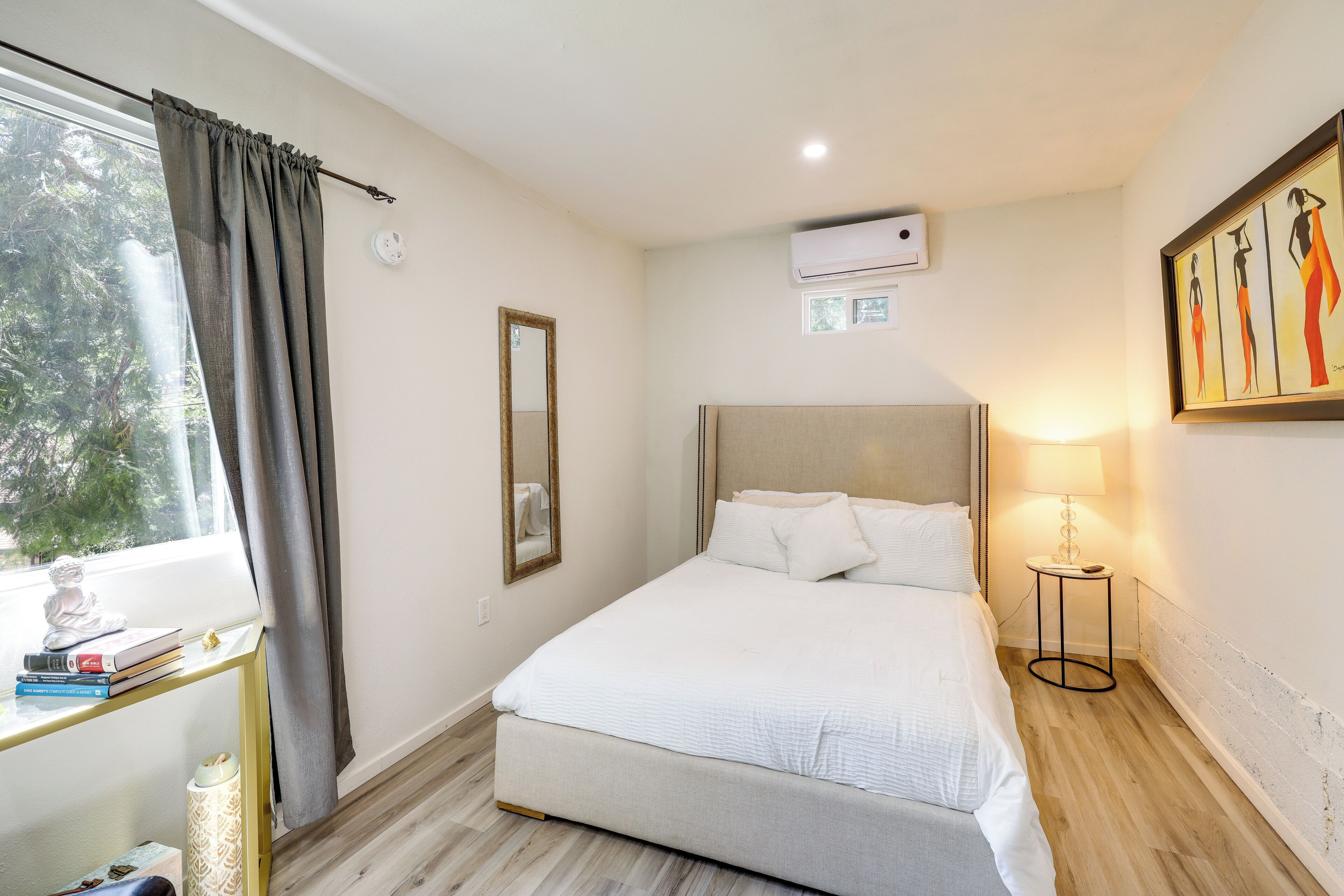 Une chambre à coucher comprenant un lit, une table de chevet, une lampe, une fenêtre avec des rideaux et un climatiseur fixé au mur.