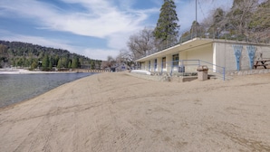 Un bâtiment situé au bord de la plage, avec une plage de sable au premier plan, un ciel dégagé et une colline boisée en arrière-plan.
