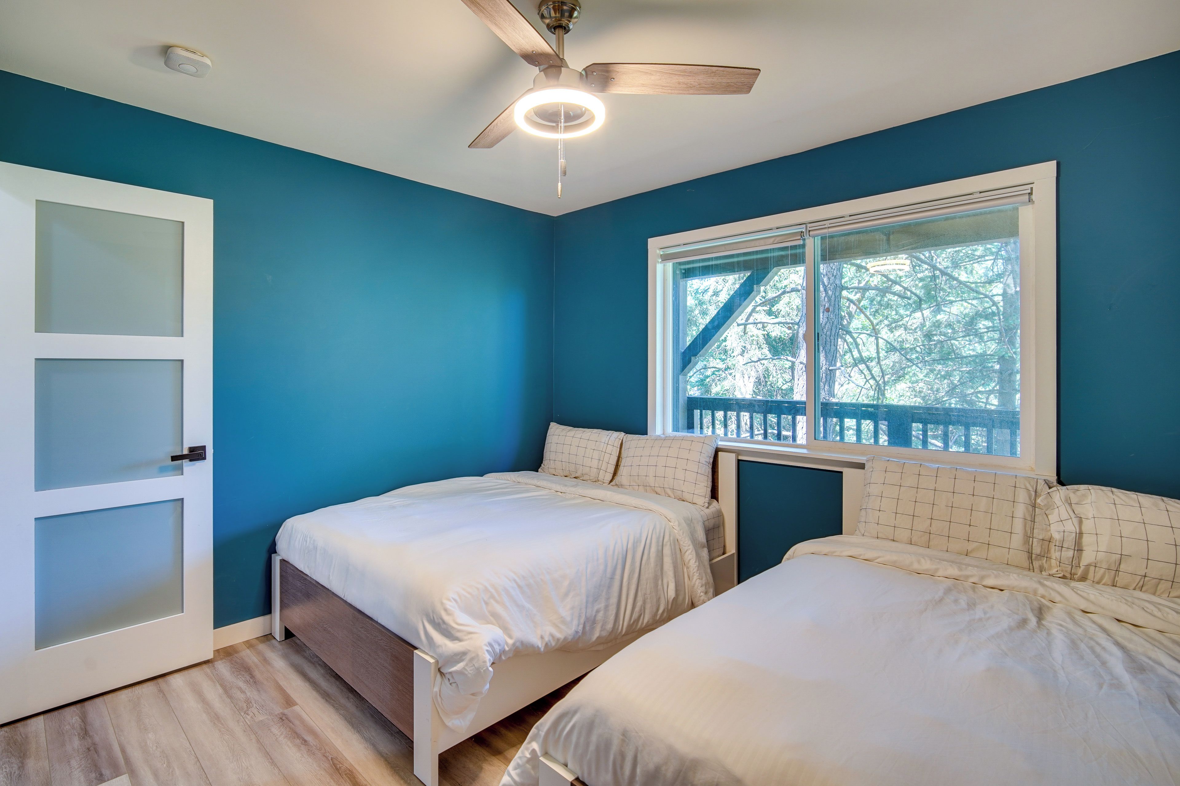 A bedroom with two beds, a ceiling fan, a window with a view of trees, and a white door with frosted glass panels.