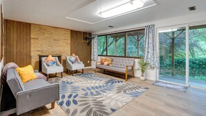 A modern living room with a grey sofa, two armchairs, a wooden accent wall, a large window with a view of greenery, and a ceiling light fixture. - Expansive Lake Forest Park Home w/ Private Deck! (Lake Forest Park)