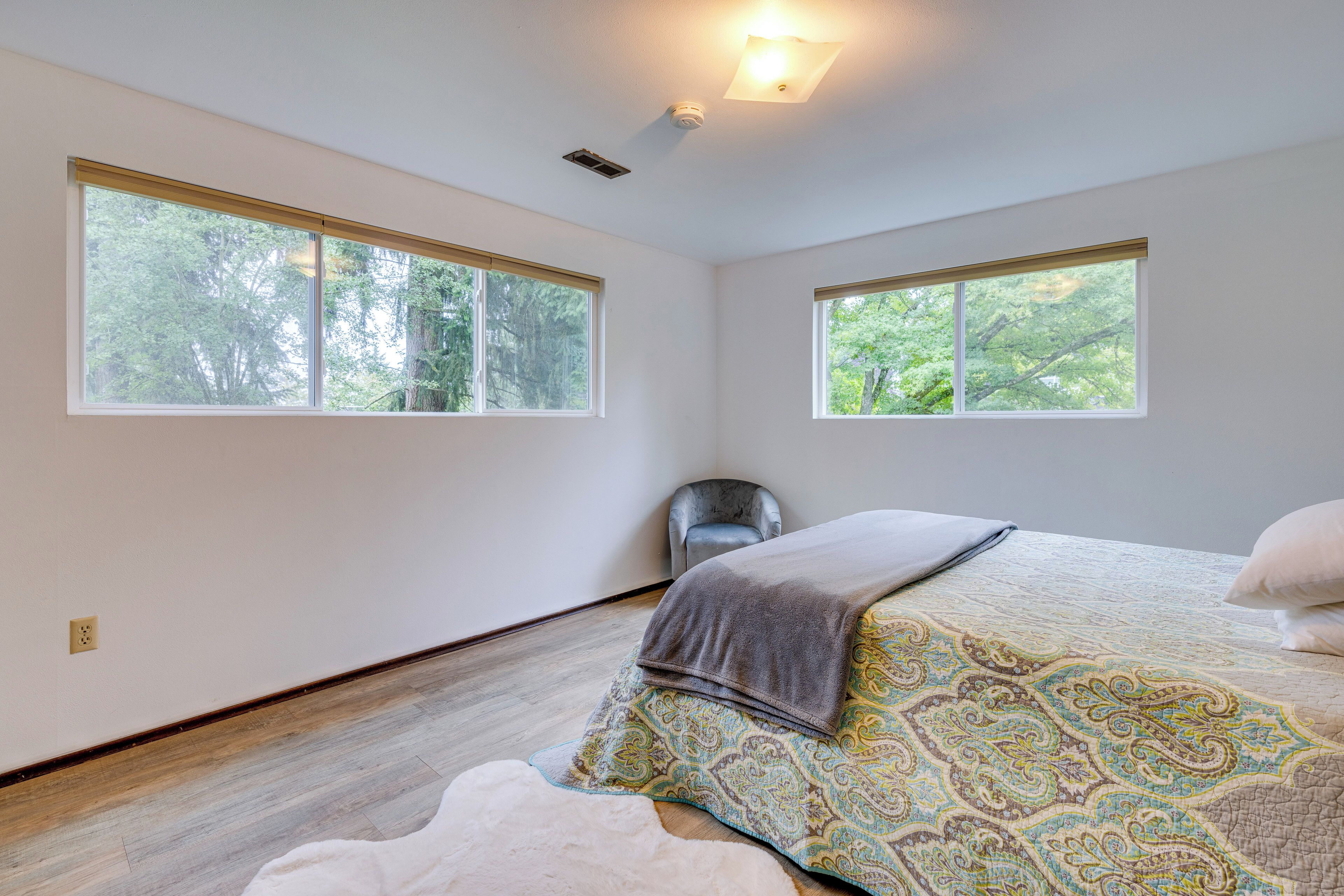A bedroom with two large windows, a bed with a patterned bedspread, a chair, and a white rug.