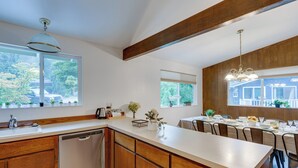A modern kitchen with wooden cabinets, a dishwasher, and a dining area with a table set for four. - Expansive Lake Forest Park Home w/ Private Deck! (Lake Forest Park)