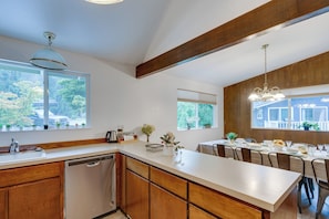 A modern kitchen with wooden cabinets, a dishwasher, and a dining area with a table set for four. - Expansive Lake Forest Park Home w/ Private Deck! (Lake Forest Park)