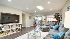 A modern living room with a flat-screen TV, glass coffee table, grey sofa, and a dining area in the background.