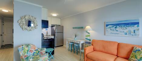 A living room with a sofa, a chair, a dining table with stools, a refrigerator, and a ceiling fan.