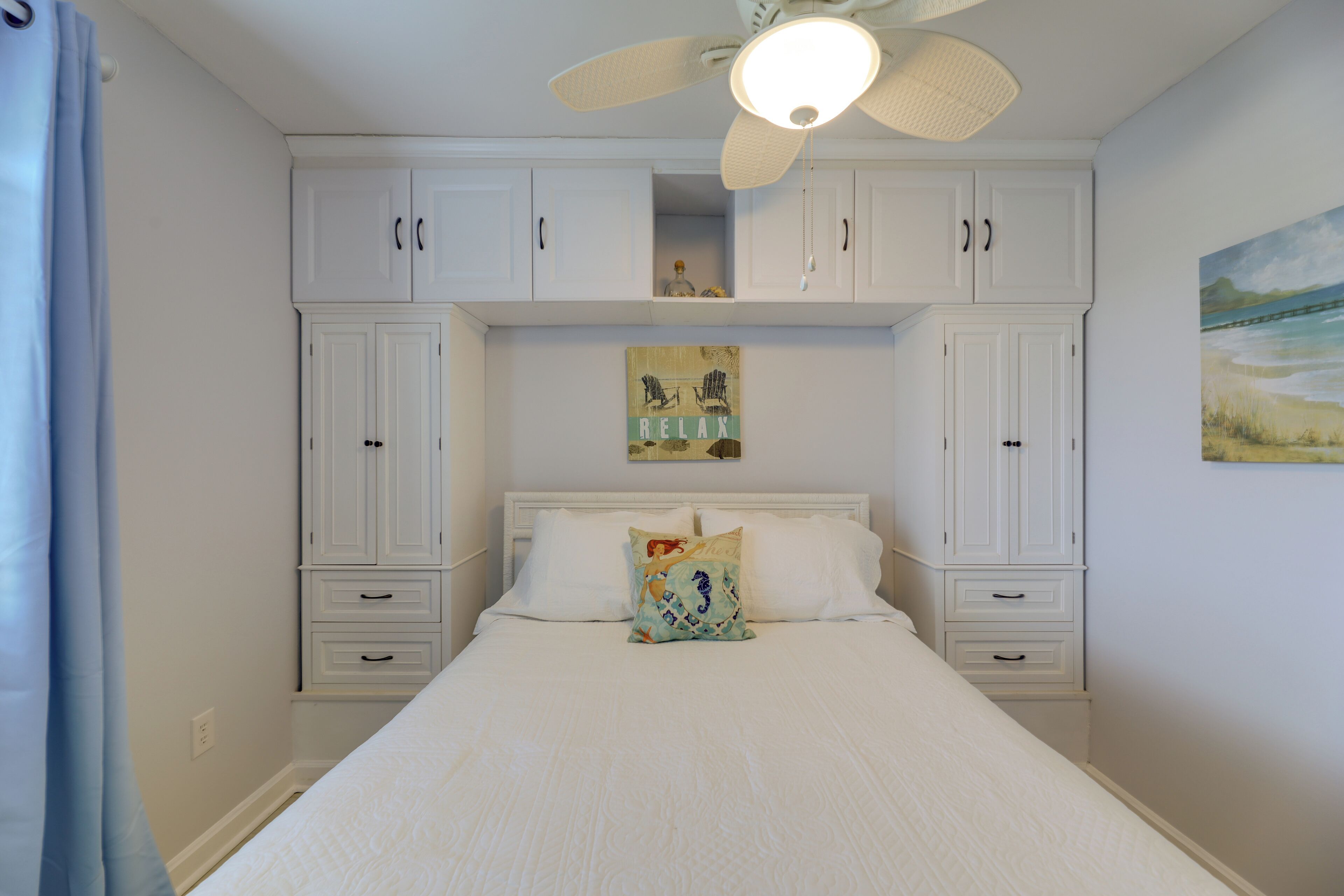 A bedroom with a bed, white cabinets, a ceiling fan, and a painting on the wall.