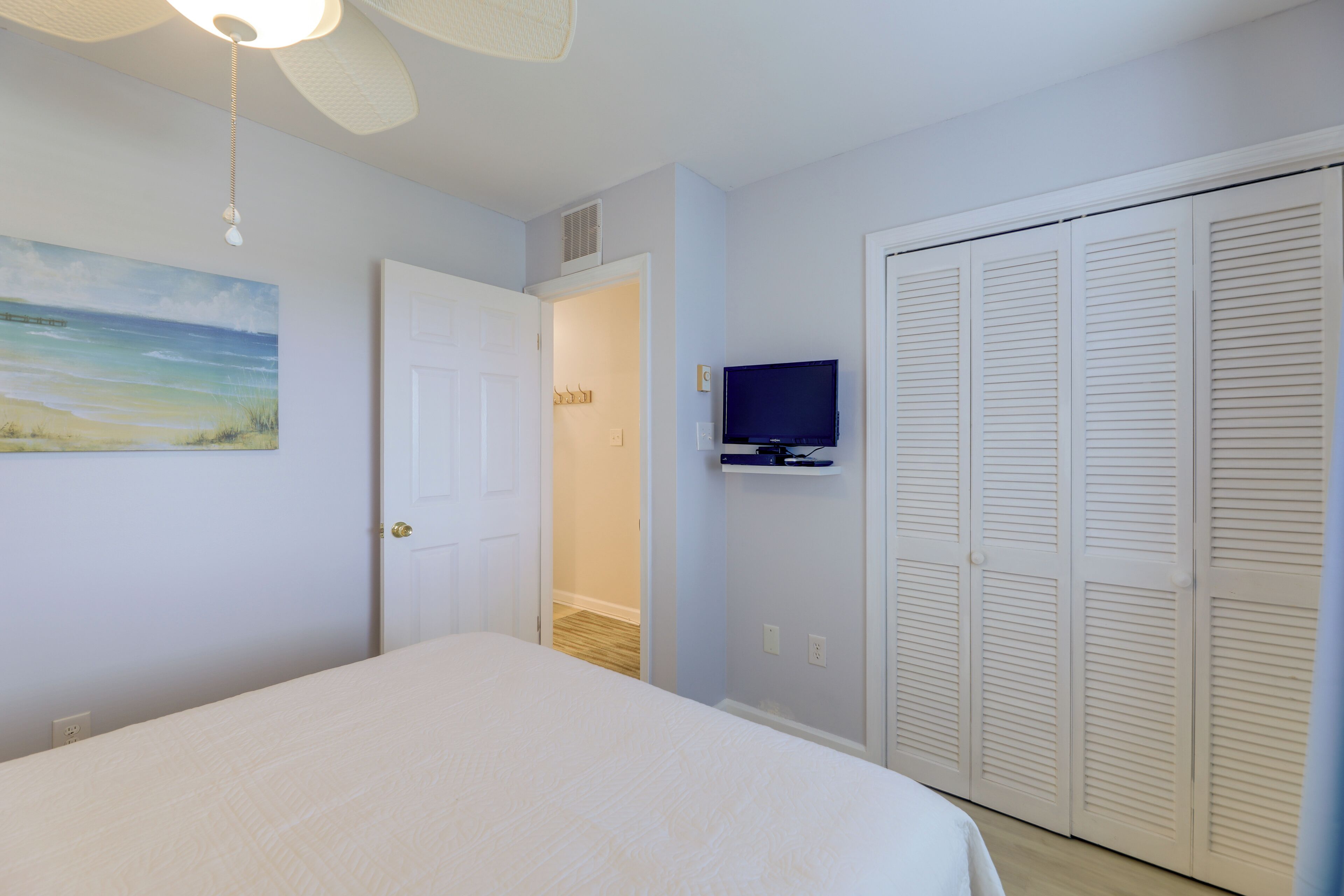 A bedroom with a bed, a ceiling fan, a TV mounted on the wall, and a closet with louvered doors.