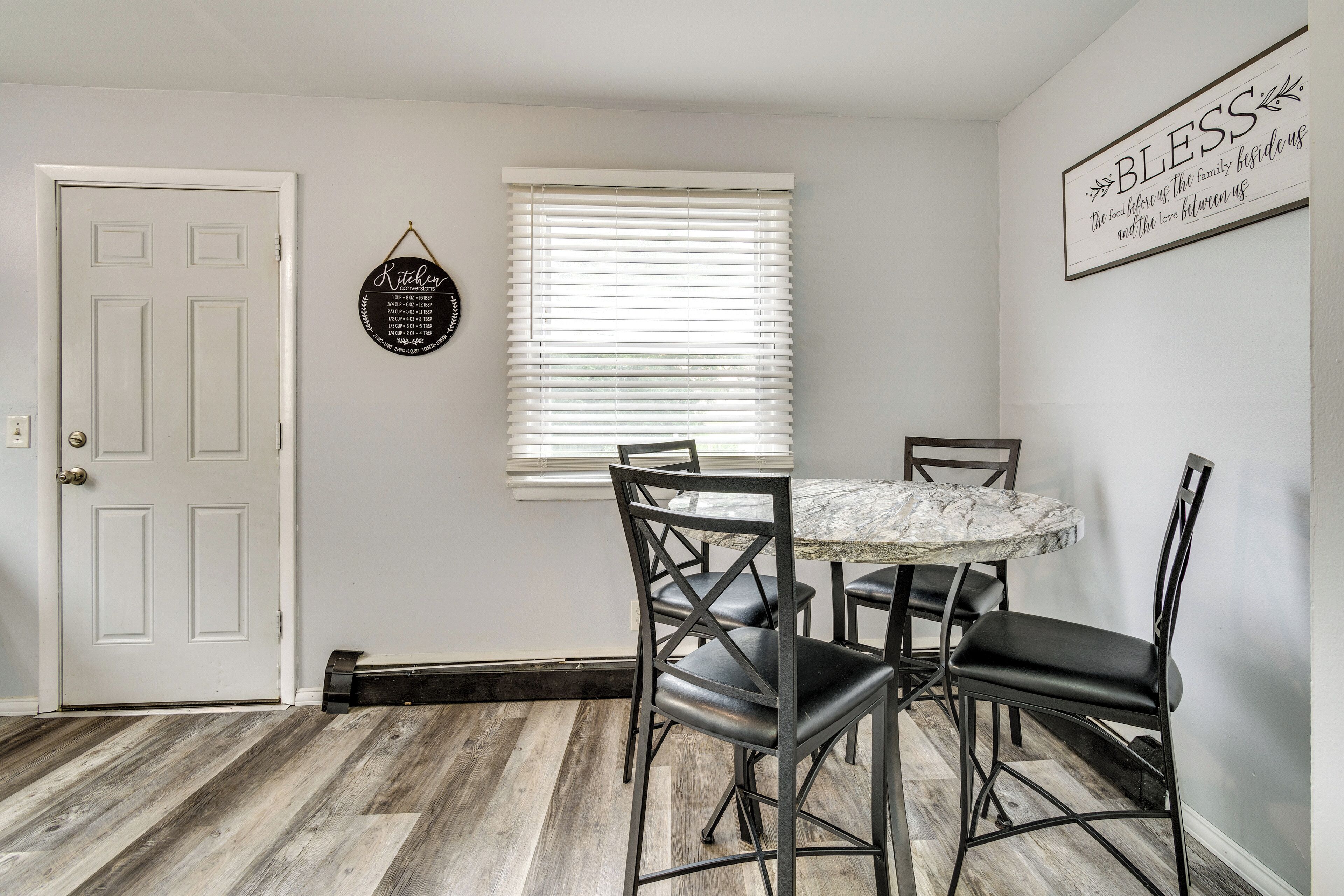A dining area with a round table and four chairs, a white door, a window with blinds, and a wall sign.