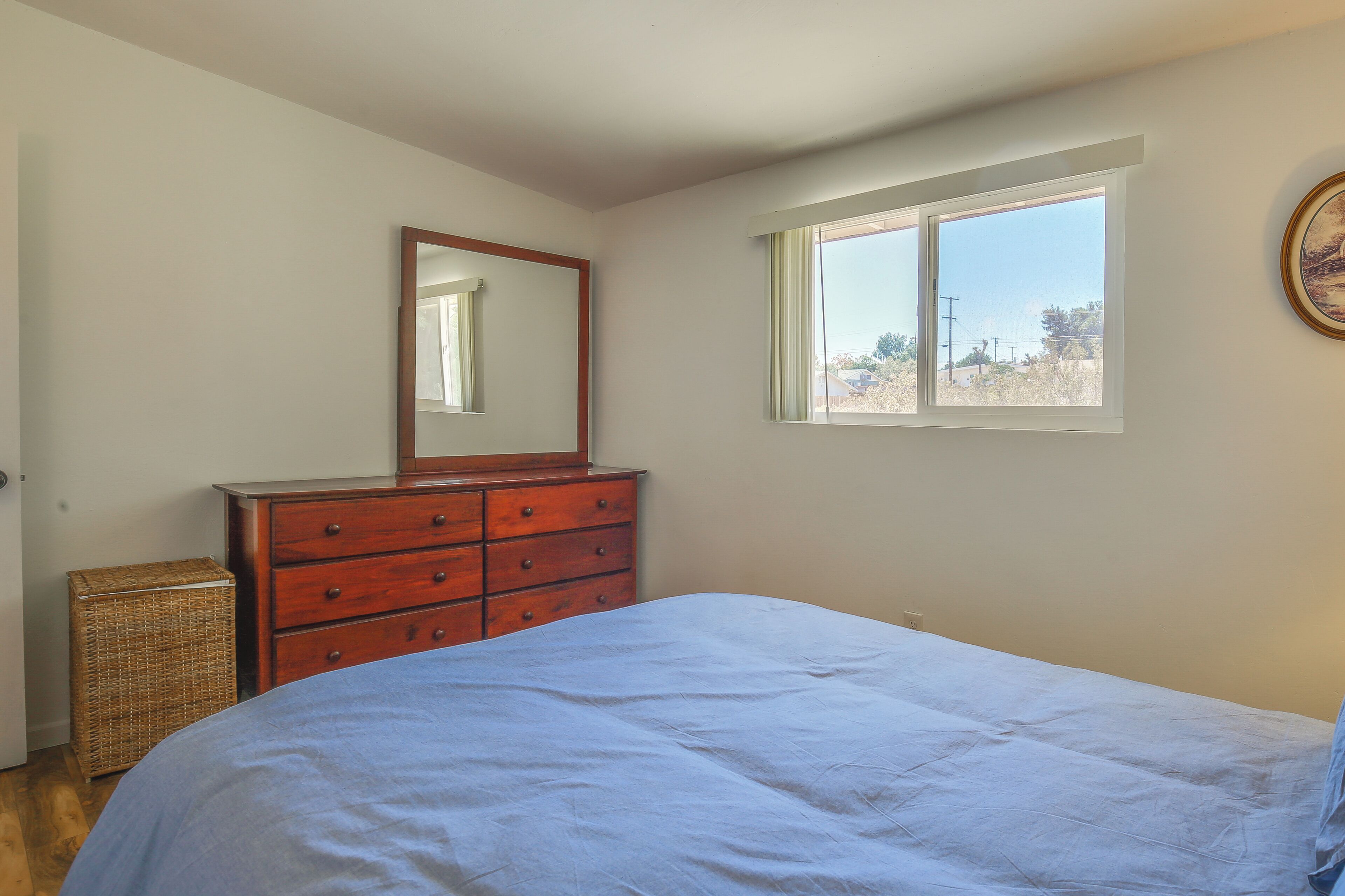 A bedroom with a bed, a dresser, a wicker basket, and a window with a view.