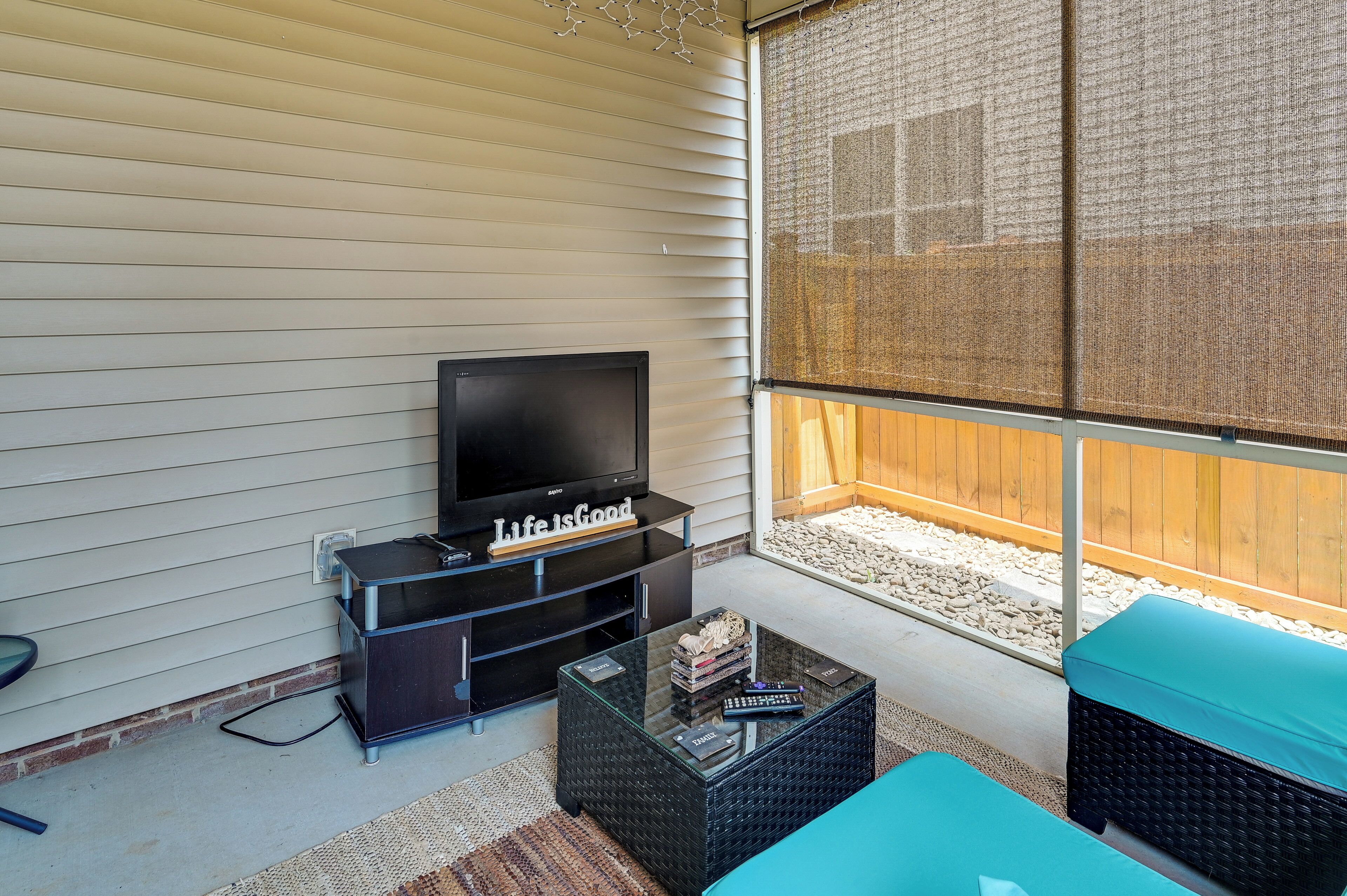 A screened-in porch with a television on a black stand, a glass coffee table, and a blue cushioned bench.