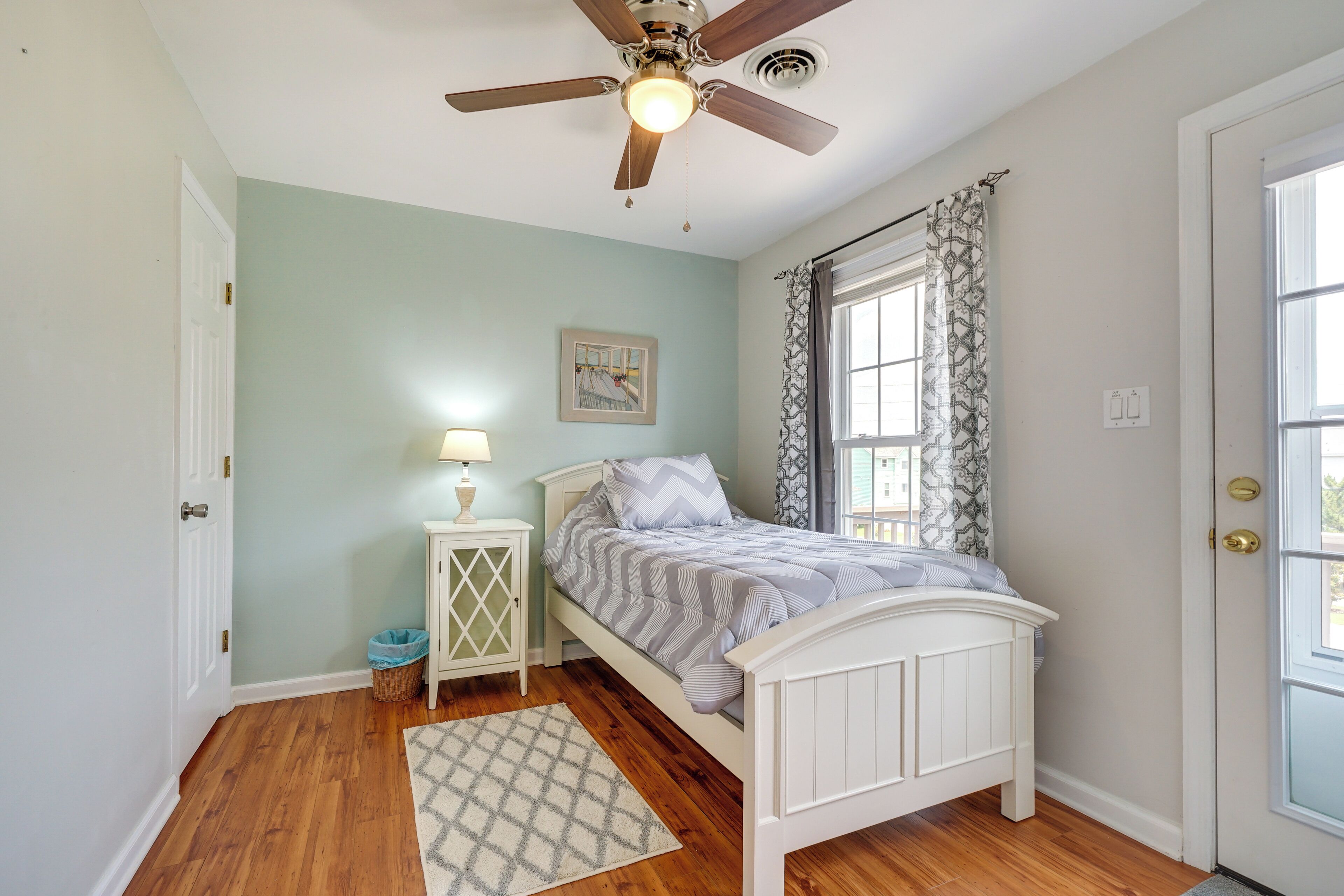 A bedroom with a bed, a ceiling fan, a nightstand with a lamp, a window with curtains, and a door.