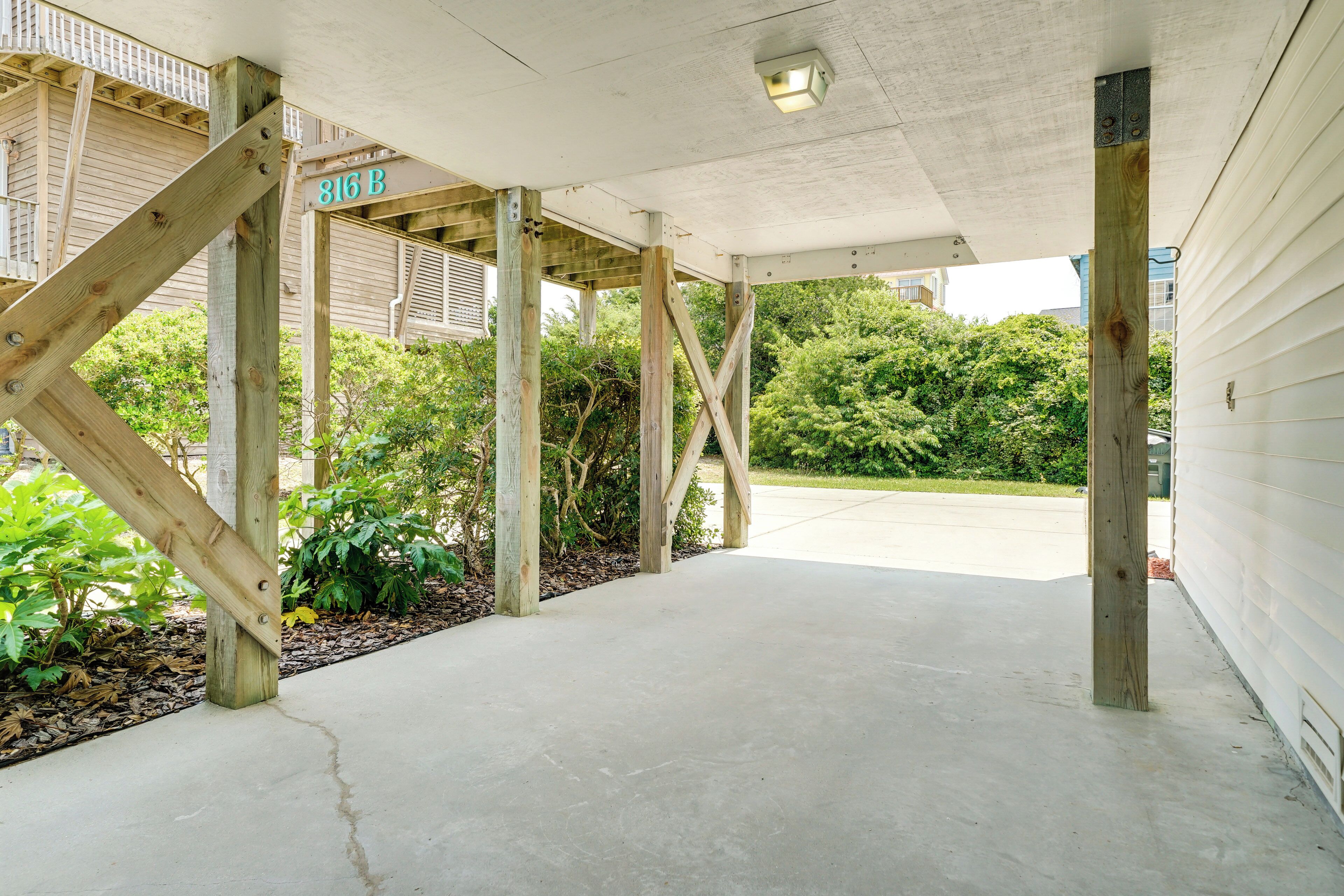 A covered outdoor area with wooden beams and a concrete floor, surrounded by greenery and a building in the background.