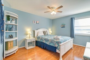 A bedroom with a bed, a nightstand, a shelf unit, a ceiling fan, and a window with curtains. - Coastal Murrells Inlet Condo With Balcony! (Murrells Inlet)
