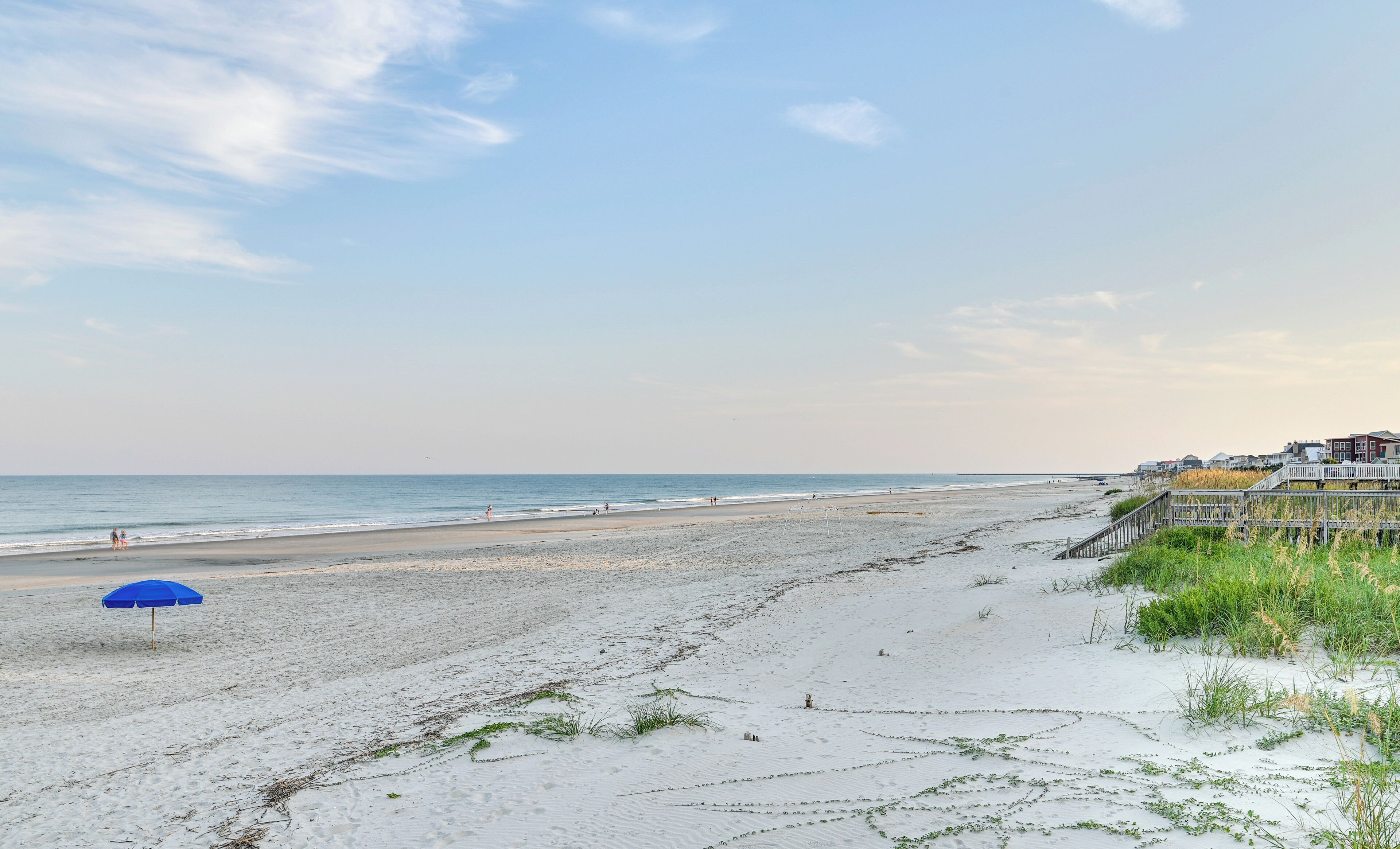 A beach with a blue umbrella, sandy shore, and a pier with buildings in the background.
