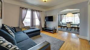 A modern living room with a sectional sofa, a flat-screen TV, and a dining area with a table and chairs. - Detroit Vacation Rental w/ Porch & Fenced Backyard (Highland Park)