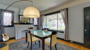 A dining area with a glass table, four chairs, a guitar, a fireplace, and a large window with curtains. - Detroit Vacation Rental w/ Porch & Fenced Backyard (Highland Park)