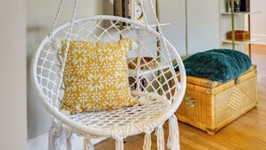 Une chaise en macramé blanc, agrémentée d’un coussin jaune à motifs, d’un plaid turquoise et d’une commode en osier.