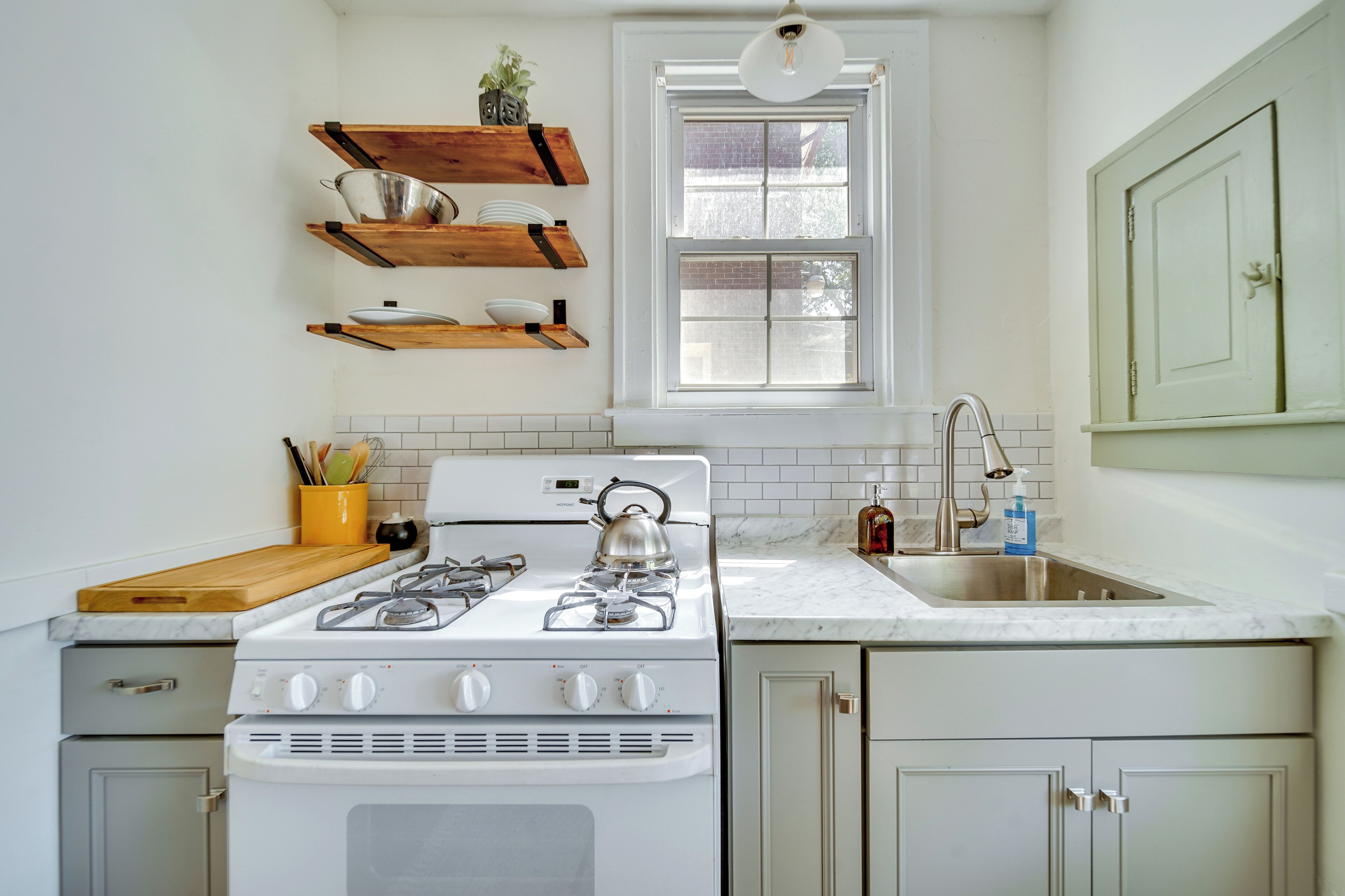 Una cocina con gabinetes blancos, estufa, pileta y estanterías de madera sobre la estufa, además de un ventanal.