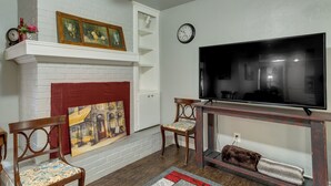 Una sala de estar con chimenea, dos sillas de madera, un televisor sobre un mueble de madera y un reloj en la pared.