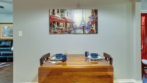 Comedor con mesa de madera preparada para cuatro personas, un cuadro enmarcado de un paisaje urbano arriba y una cortina roja a la derecha.