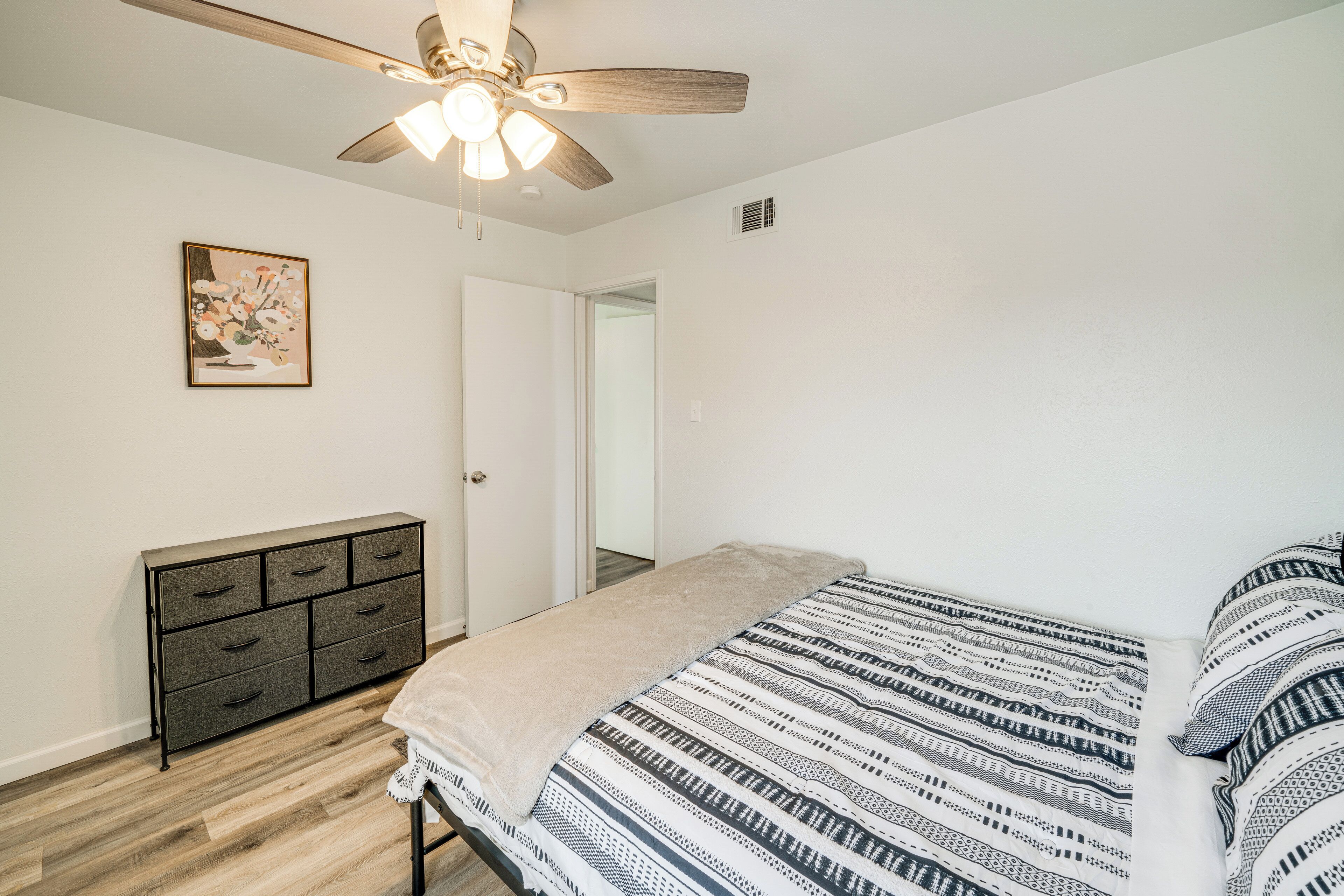 A bedroom with a bed, a dresser, a ceiling fan, and a door leading to another room.
