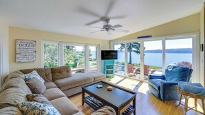A living room with a beige sectional sofa, a wooden coffee table, a blue armchair, and a view of the ocean through large sliding glass doors.
