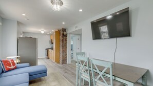 A modern living room with a blue sofa, a dining table with chairs, a TV mounted on the wall, and a kitchen area in the background. - Sunny Seaside Heights Retreat: Walkable Location! (Seaside Heights)