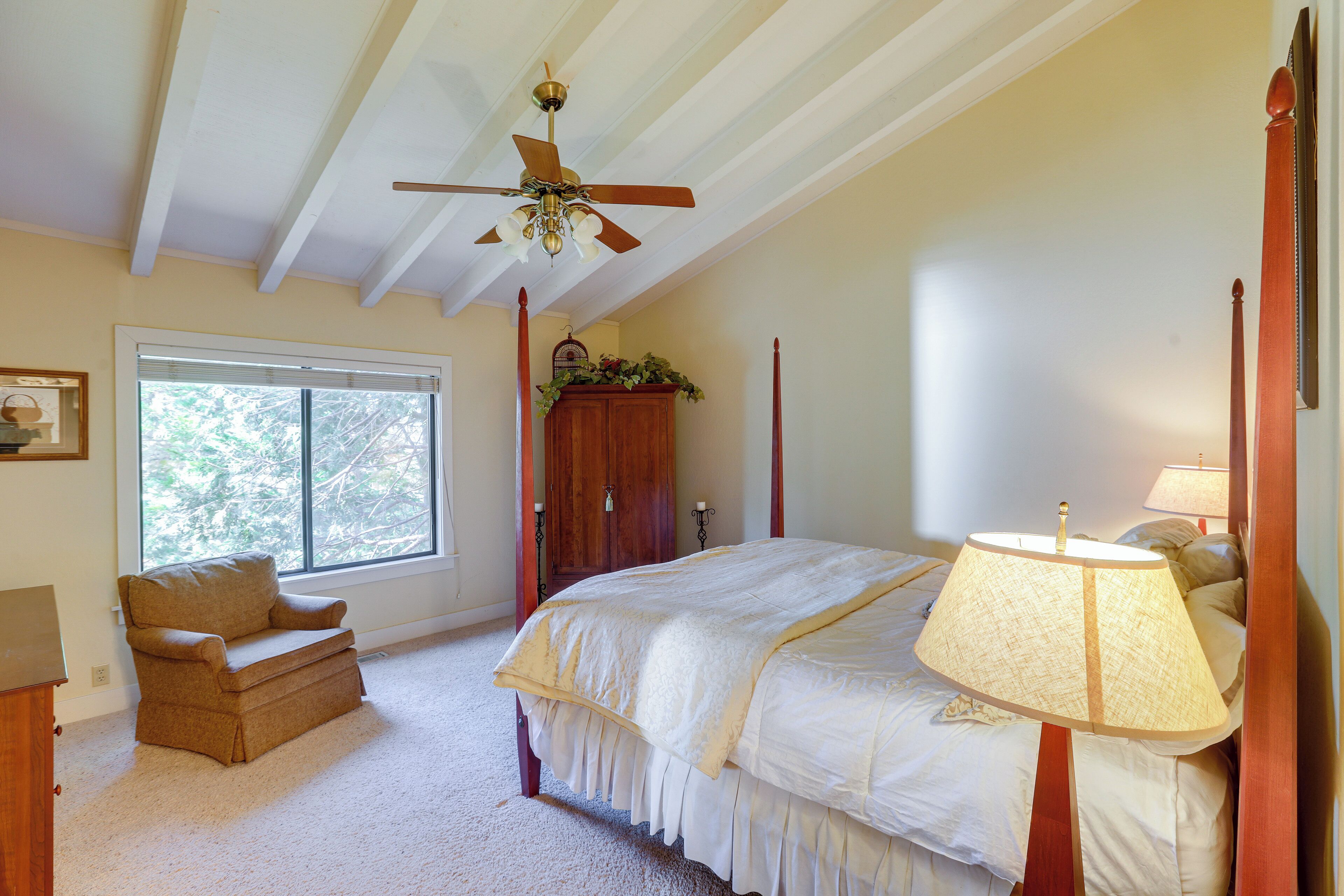 Une chambre à coucher avec un grand lit, un ventilateur de plafond, une fenêtre, une armoire en bois et un petit fauteuil.